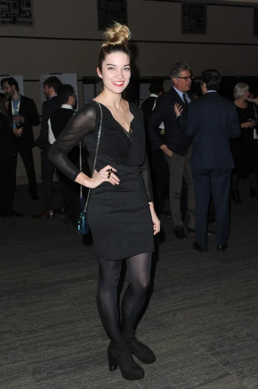 Annie Murphy smiling with a top knot in a fitted black dress with sheer sleeves, black opaque tights and platform shoes