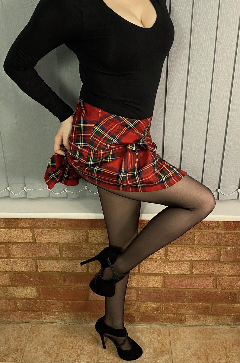 Woman in a black top and red tartan mini skirt standing on one leg against a brick wall in sheer black pantyhose and black Mary Jane heels