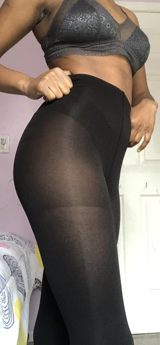 Woman with dark skin standing in a bedroom with a light purple wall in a grey lace bralette pulling black tights up over her hips with both hands at the waistband