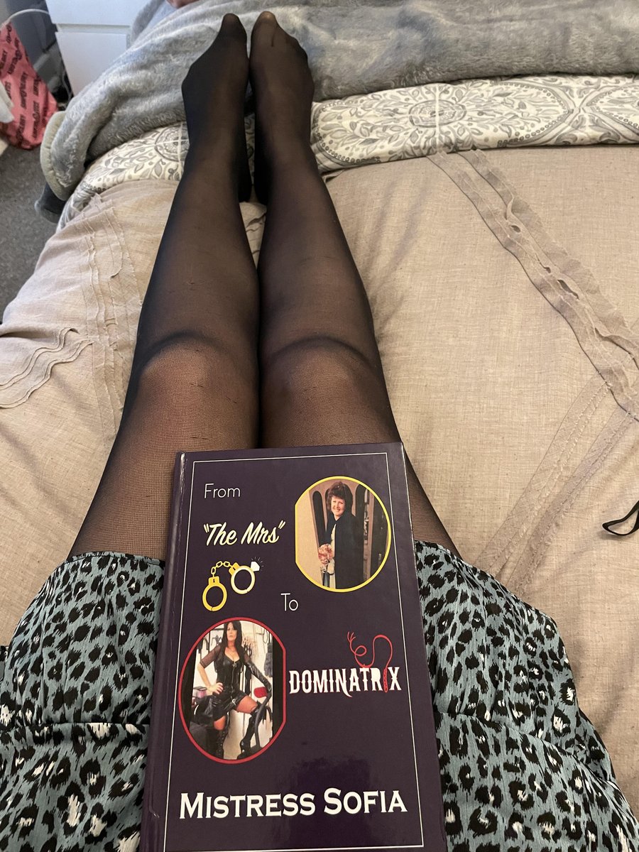 Legs in sheer black pantyhose on a bed with a book titled Dominatrix