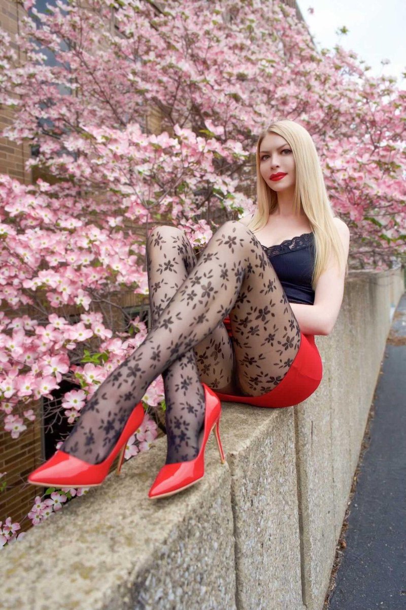 Blonde woman with bright red lipstick in a black sleeveless top with lace trim and a bright red skirt sitting on a rough concrete ledge with crossed legs in sheer black tights with a repeating flower pattern and glossy red pointed stilettos