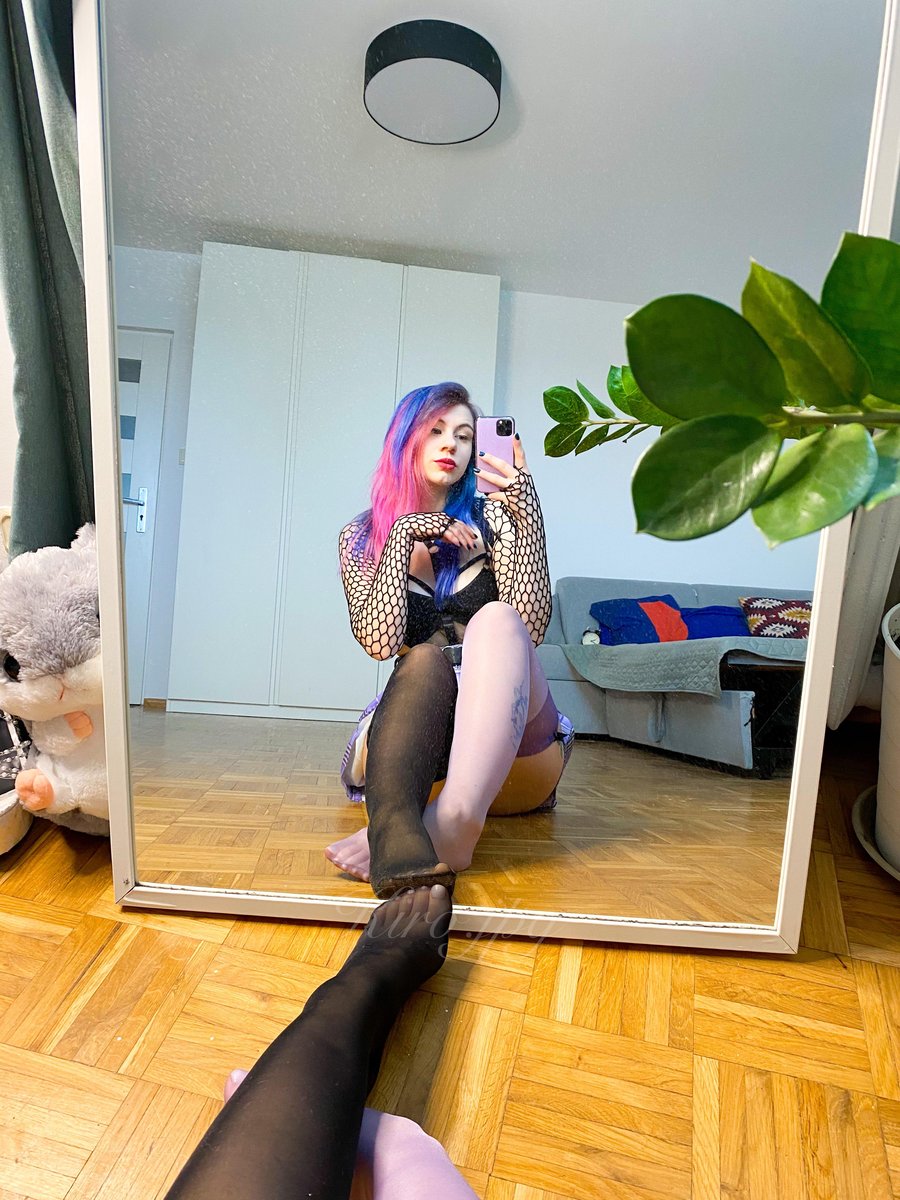 Woman with pink and blue dyed shoulder-length hair in a black cutout top with long fishnet sleeves and a short lilac skirt with one leg extended in sheer black pantyhose holding a phone