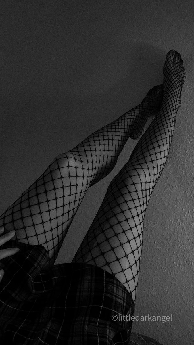 Black and white photo of legs raised against a textured wall with ankles crossed in black fishnet tights and a dark plaid garment with a hand resting near the hip