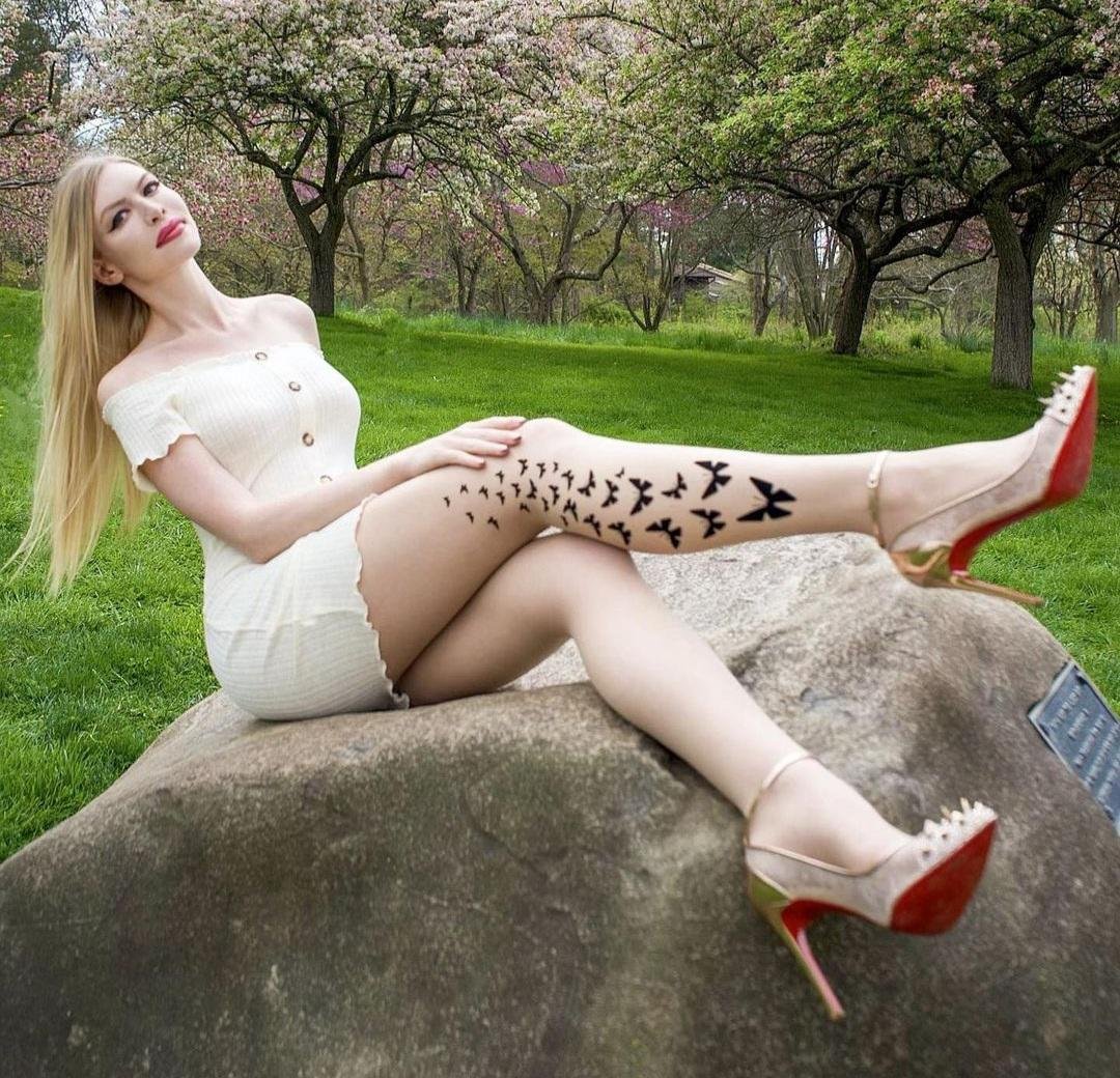 Blonde woman in a cream dress sitting on a large rock in a blossoming park wearing butterfly print pantyhose and studded red sole heels