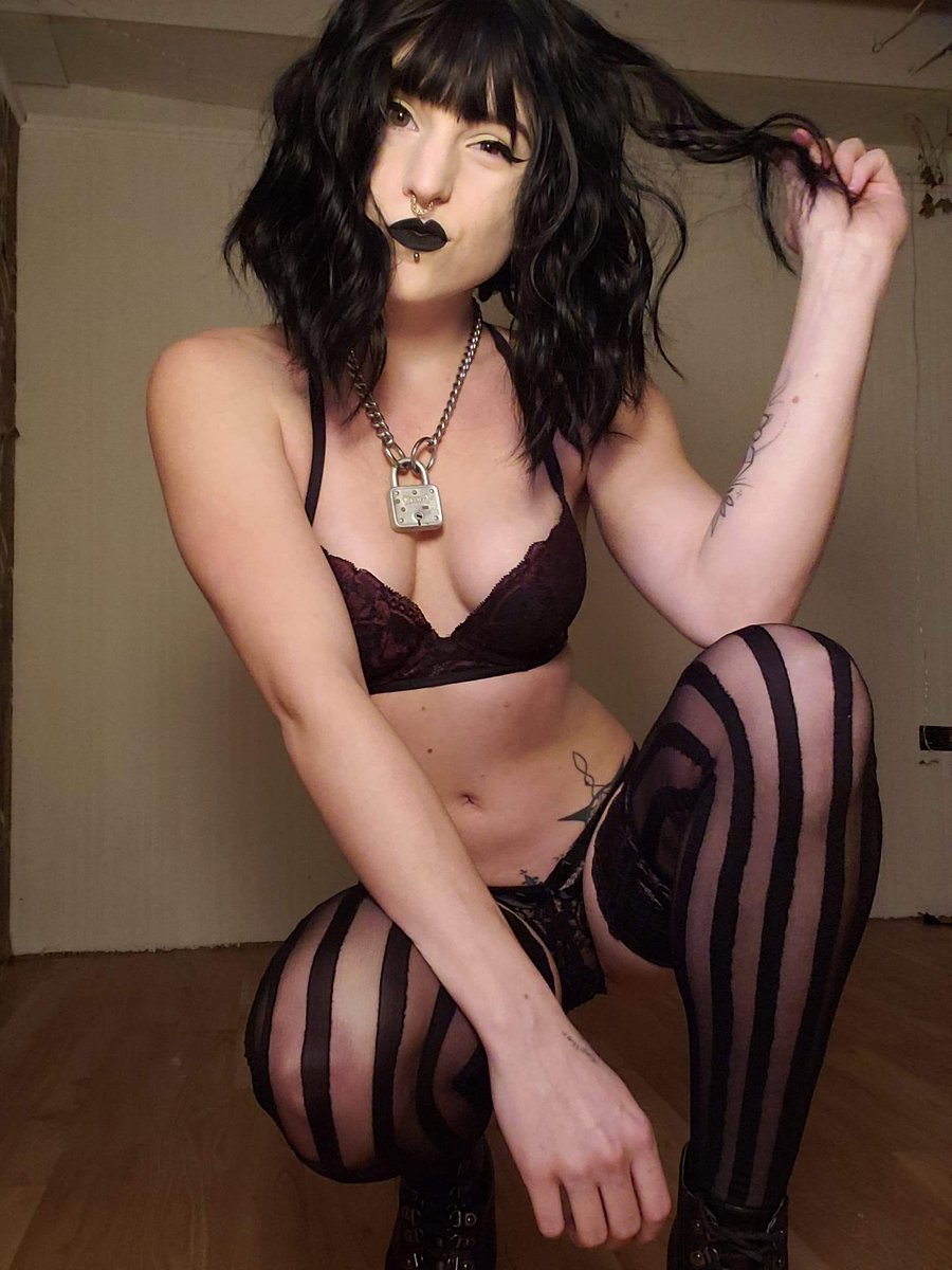 Goth girl with short dark hair and black lipstick in a bra squatting in purple and black striped stockings and black boots