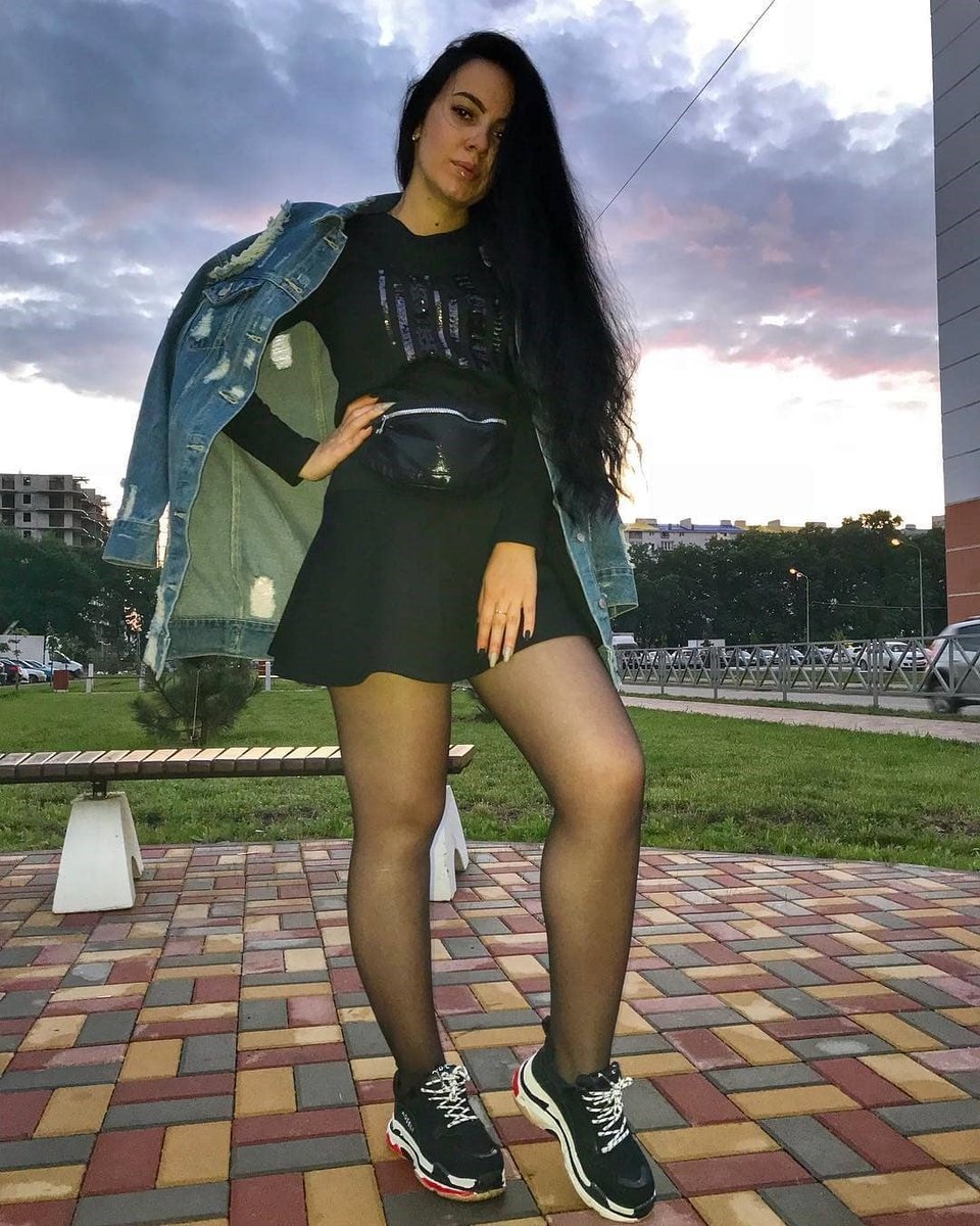Woman with very long dark hair in a denim jacket and black skirt posing in sheer black pantyhose and sneakers at sunset
