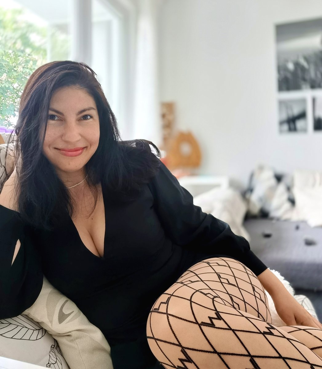 Mature woman with dark hair in a black top smiling on a sofa with one leg in white fishnet tights
