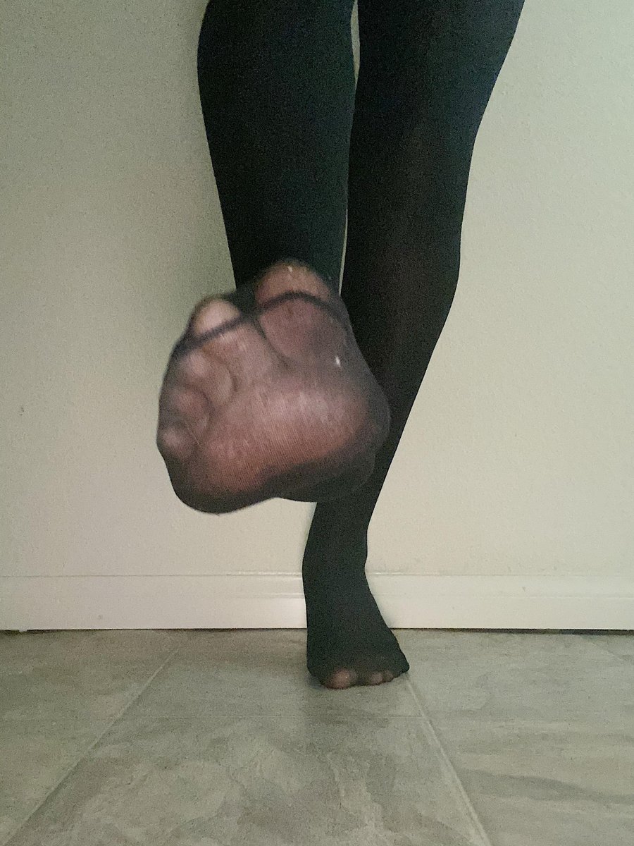 Foot in black pantyhose lifted showing a worn toe against grey tiles and a wall