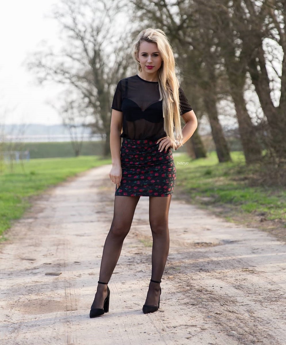 Long-haired blonde in a sheer black top and red print mini skirt standing on a dirt road in black pantyhose and heels