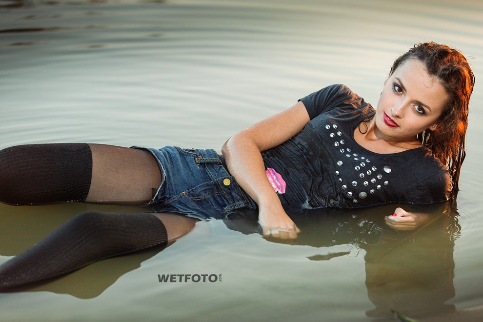 Brunette lying in shallow water in a black studded t-shirt, denim shorts and black thigh-high stockings at sunset