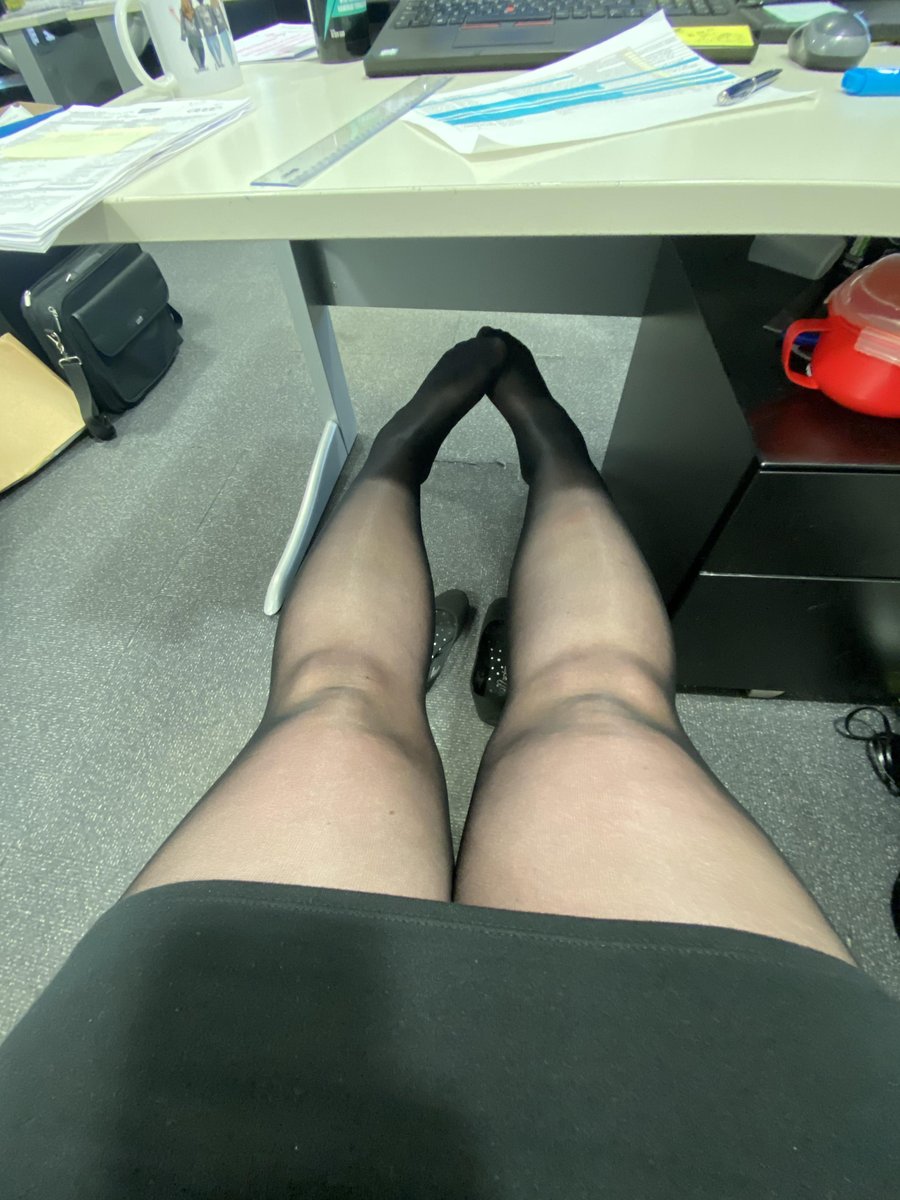 Woman seated under a desk with legs extended and ankles crossed in a black skirt and sheer black pantyhose with black pointed-toe heels with studded details in an office with a laptop, papers, ruler and mug