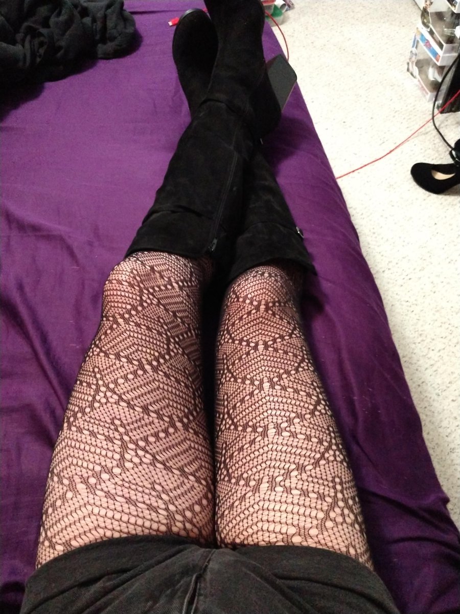 Legs lying on a purple bed in pink patterned fishnet tights and black suede thigh-high boots with a black mini skirt