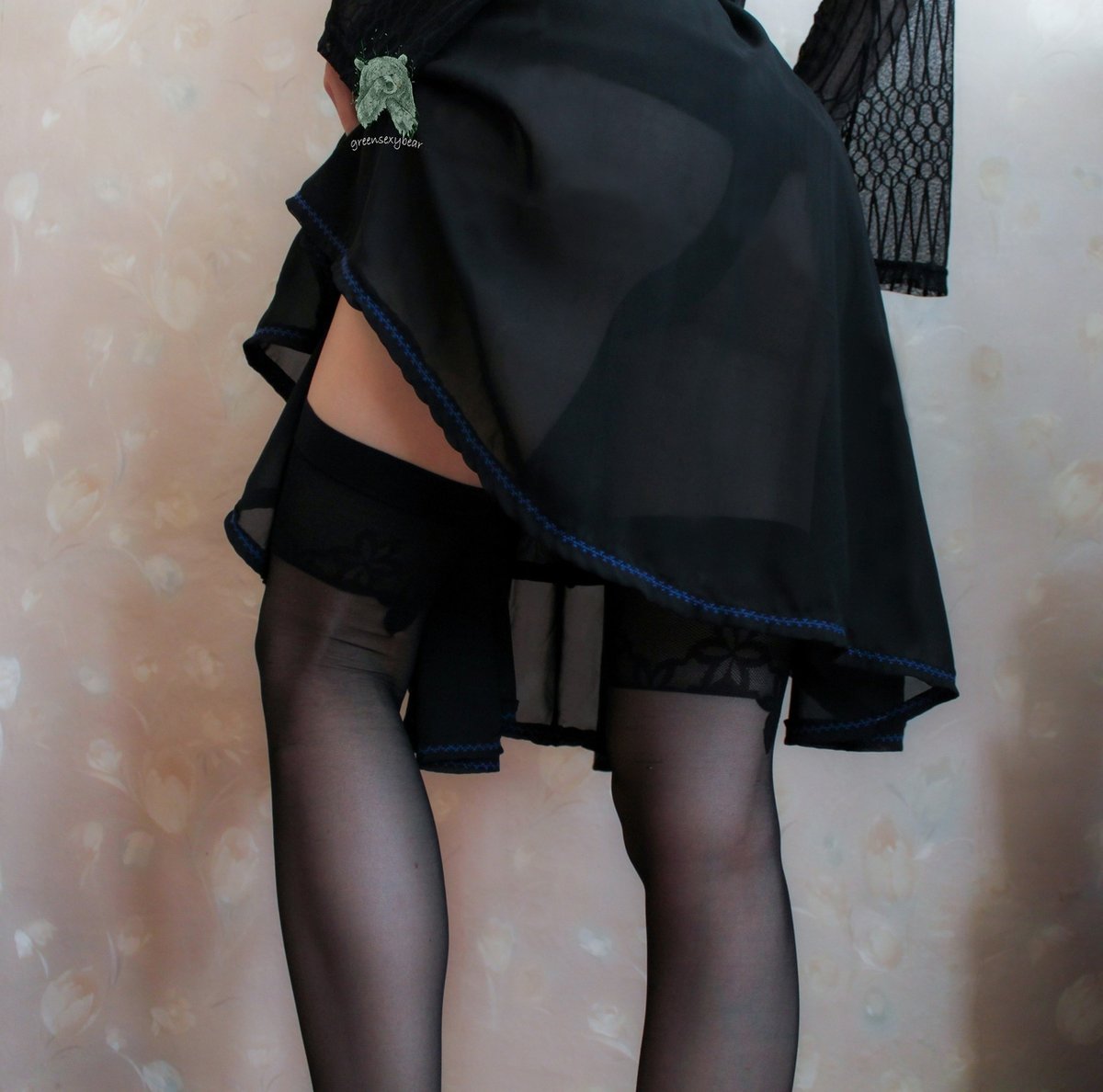 Woman in a sheer black skirt with blue trim and a black lace top standing in front of a floral wall in black stockings with thick tops and a suspender strap