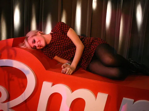 Pixie Lott with platinum hair lying on a red sofa in a red tartan mini dress and black opaque tights