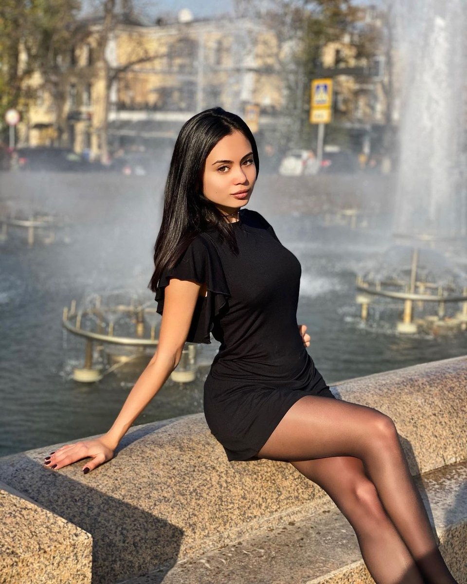 Dark-haired woman in a black dress sitting on a fountain edge in black pantyhose