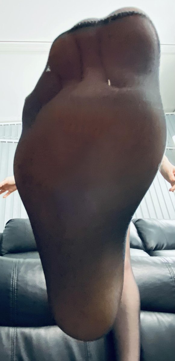 A sole in worn dark pantyhose pushed toward the camera over a black leather sofa
