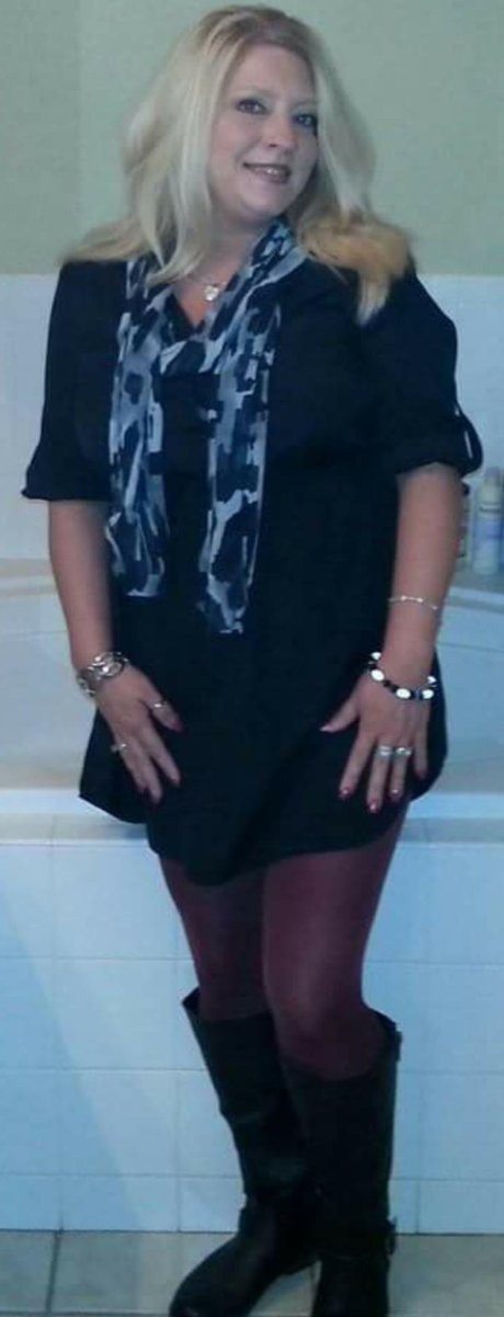 Mature blonde woman in a navy dress and scarf standing by a bathtub in burgundy tights and black knee-high boots