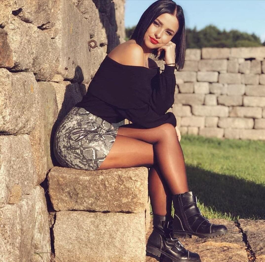 Brunette with short dark hair in a black off-shoulder top and snakeskin pattern skirt sitting on a stone wall in sheer pantyhose and black boots