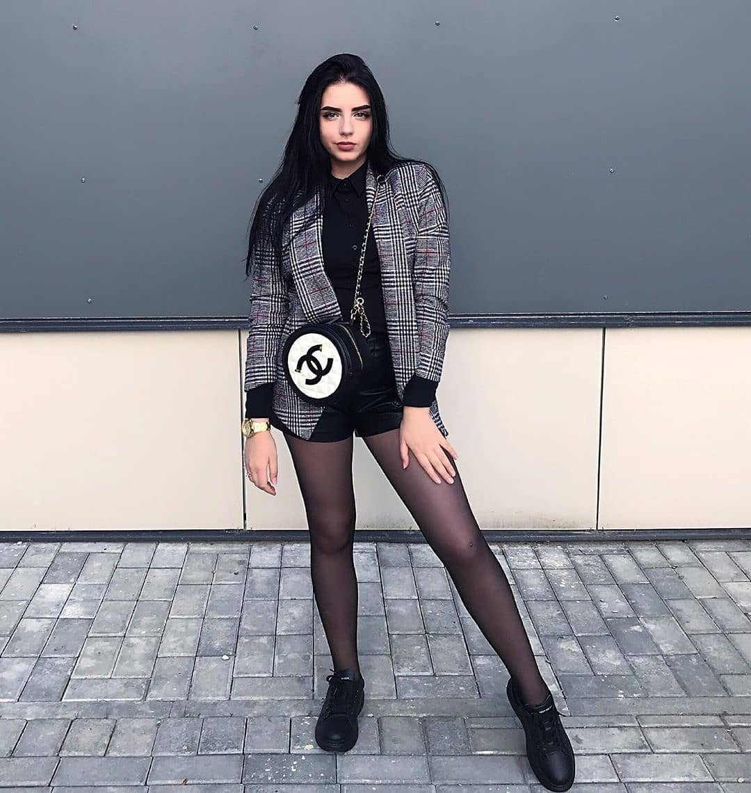 Dark-haired woman in a plaid blazer and black shorts standing with legs apart in sheer black pantyhose and sneakers