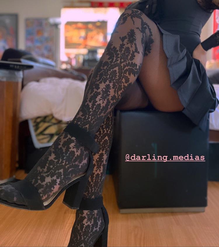 Legs in black floral lace tights and black ankle strap block heels with a grey pleated skirt on a footstool